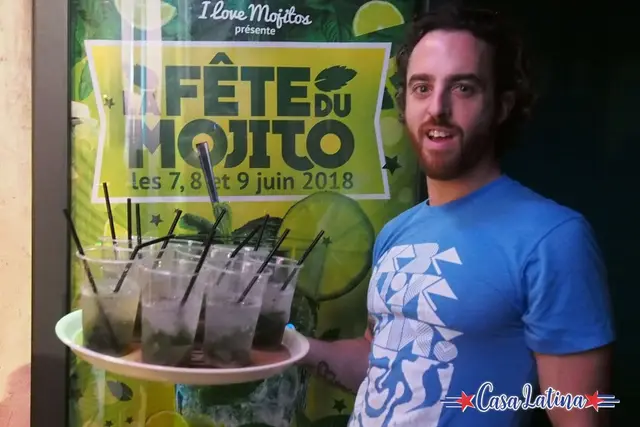 Un serveur présente un plateau de mojitos devant l'affiche de la Fête du Mojito 2018